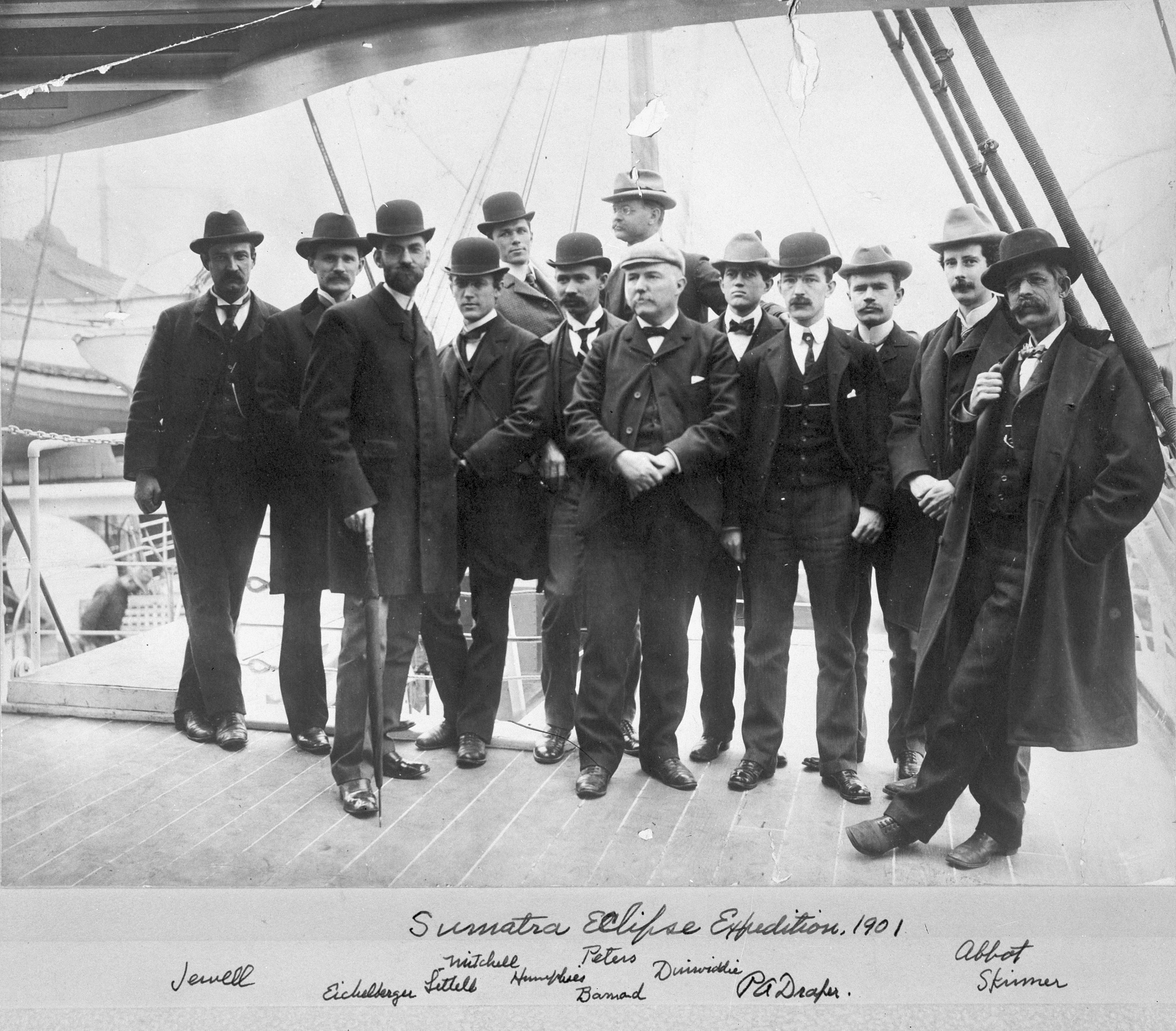 Members of the SAO's Sumatra Eclipse Expedition in 1901 pose for a photo on a deck of a ship in harbor, all wearing suits, ties, and hats.
